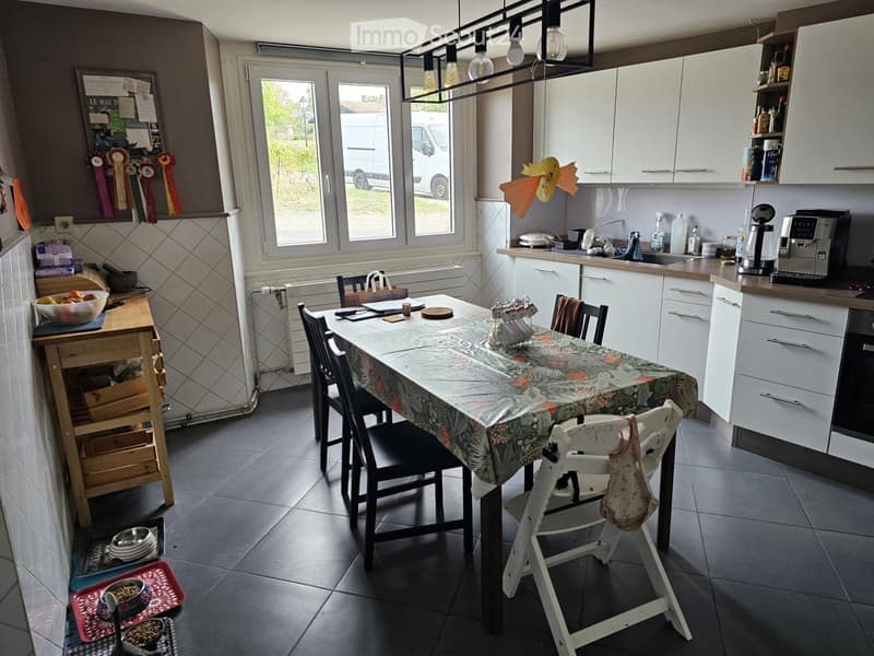 Bright kitchen with white cabinets, a central dining table with chairs, and various appliances including a coffee machine and a sink.