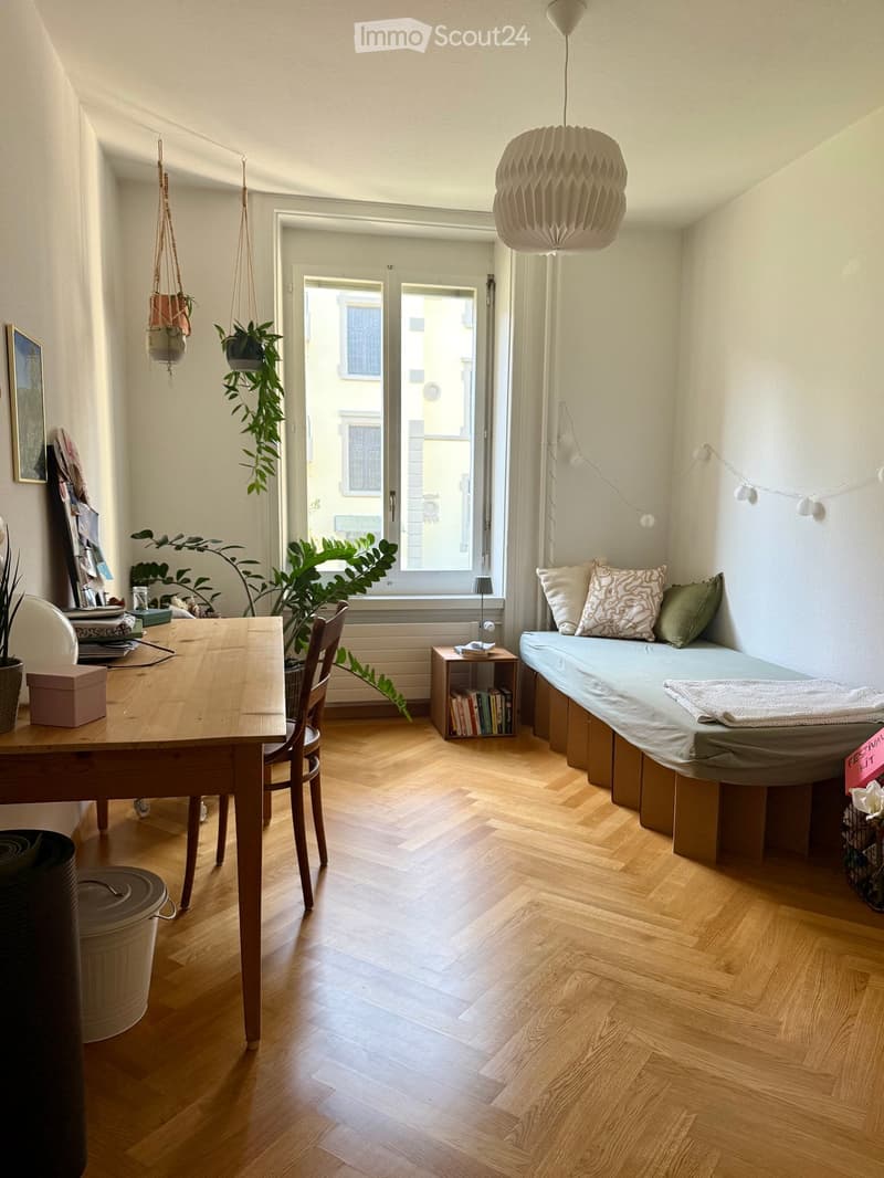 A cozy, well-lit room with wooden flooring, a bed with pillows and a blanket, a wooden table with chairs, potted plants, a bookshelf, and a hanging lamp.