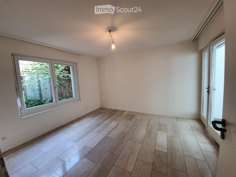 Empty room with light-colored tiled floor, white walls, and large windows showing greenery outside. No furniture or equipment present.