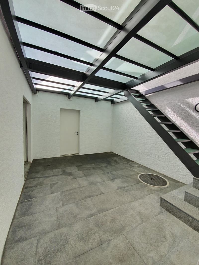Modern hallway with a glass ceiling, tiled floor, and a staircase leading upwards. Features a white door and a metal manhole cover on the floor.
