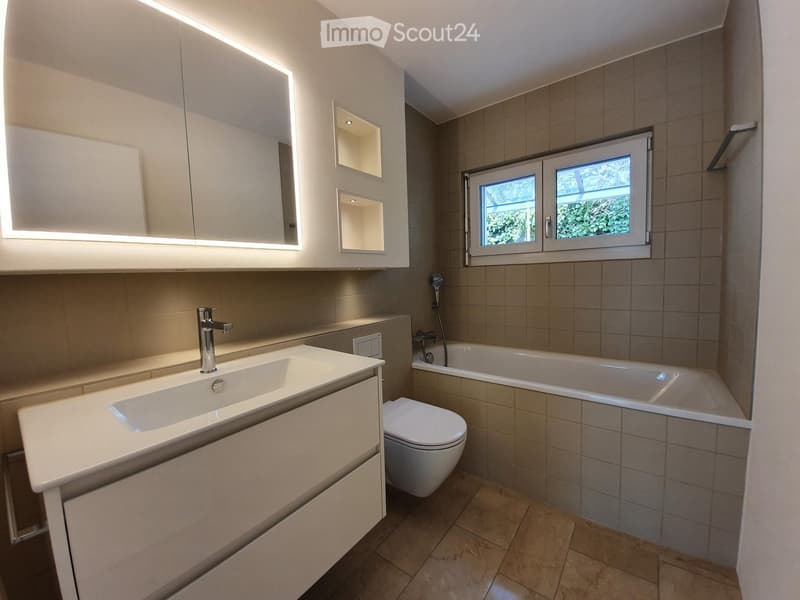 Modern bathroom with a white sink, mirror, toilet, bathtub, and built-in cabinets. Features a window with a view of greenery outside.