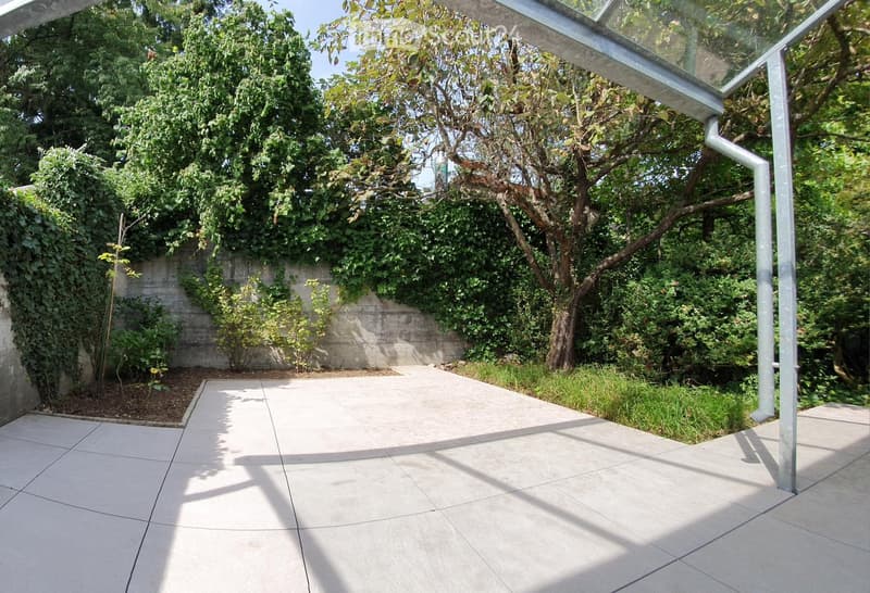 A modern outdoor patio with large tiled flooring, surrounded by lush greenery and trees, featuring a glass-roofed structure on one side.