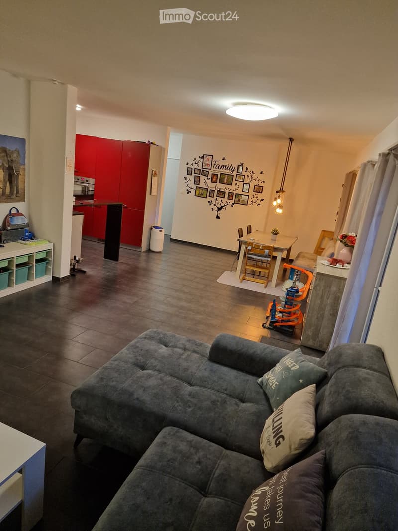 A cozy living room with a dark grey sofa, wooden dining table, chairs, and a small kitchen area with red cabinets and a white shelf. The room features a decorative wall with framed photos and a tree design.