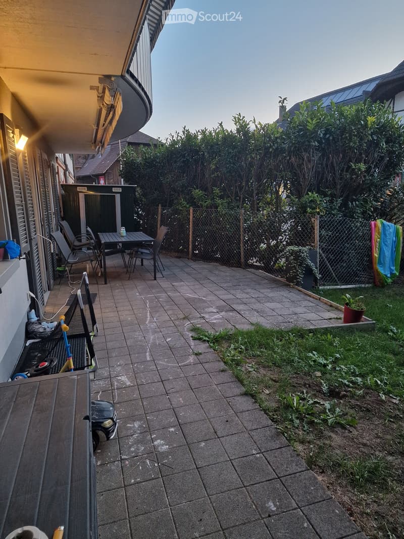 Outdoor patio area with tiled floor, small table and chairs, fenced garden with greenery, and a small toy car.