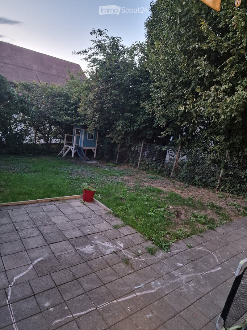 Backyard with a paved patio area, grassy lawn, small playhouse, and a red flower pot. The area is enclosed by a chain-link fence and surrounded by trees and shrubs.