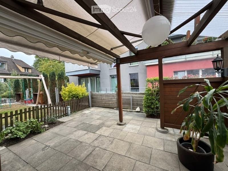 A covered patio area with wooden beams and a tiled floor, surrounded by a wooden fence and greenery, with a swing set visible in the background.