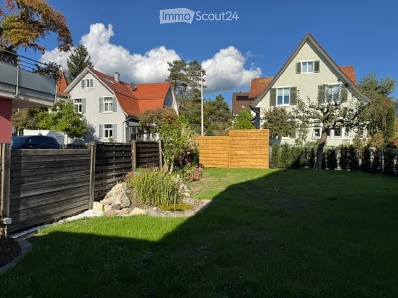 Well-maintained garden with a wooden fence, neatly trimmed grass, small shrubs, and a few trees. The background shows neighboring houses with red-tiled roofs and white walls.