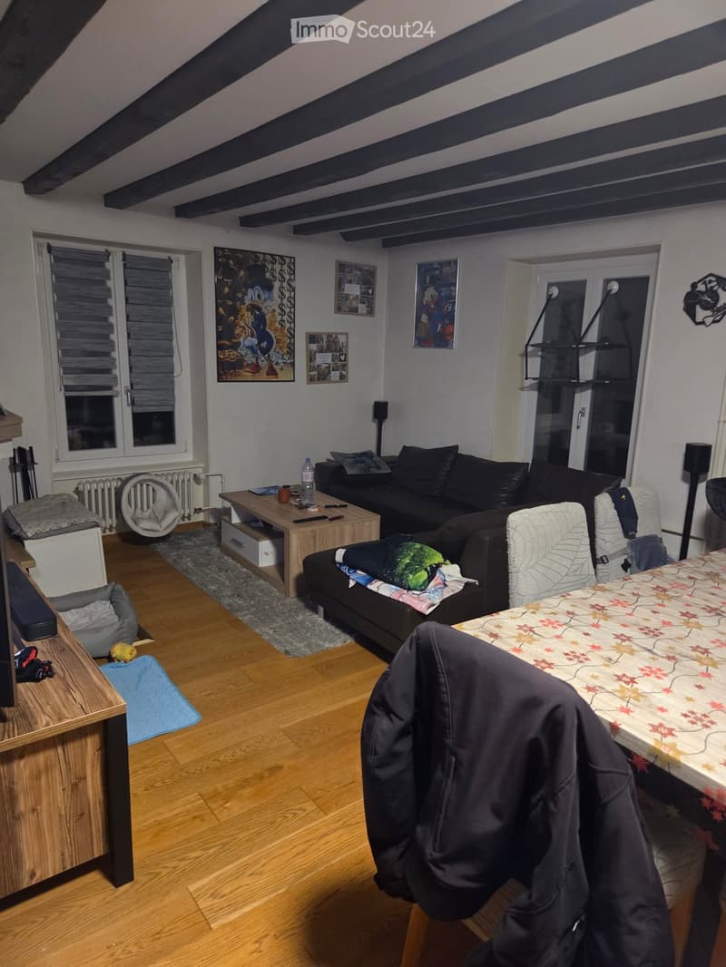 A cozy living room with wooden flooring, a black leather sofa, a wooden coffee table, a small table with a floral tablecloth, and a cat bed. The room features exposed wooden beams on the ceiling and several picture frames on the walls.