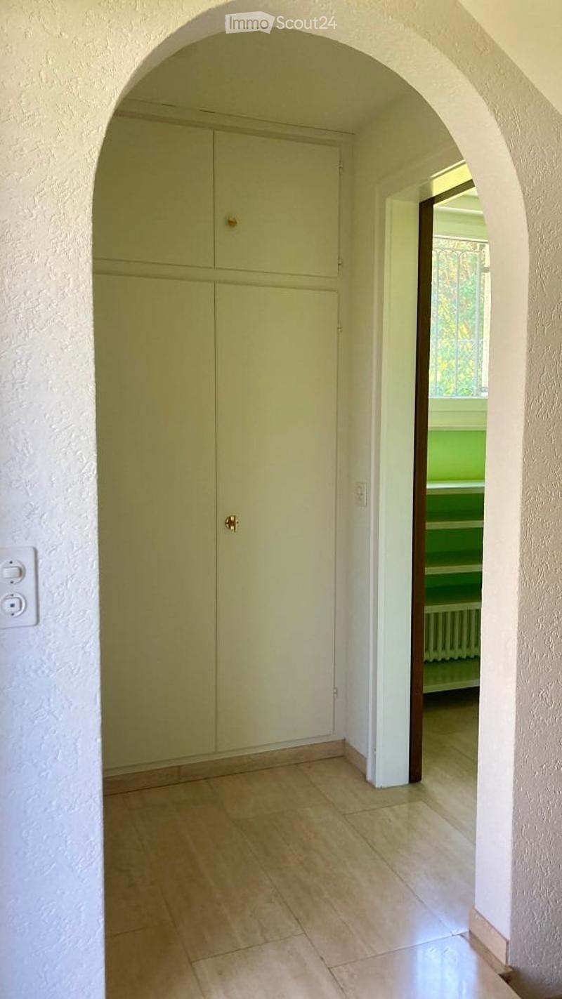 A bright hallway with a large arched doorway leading to a room. The hallway features light green built-in cabinets with gold handles, a tiled floor, and a visible light switch on the wall.