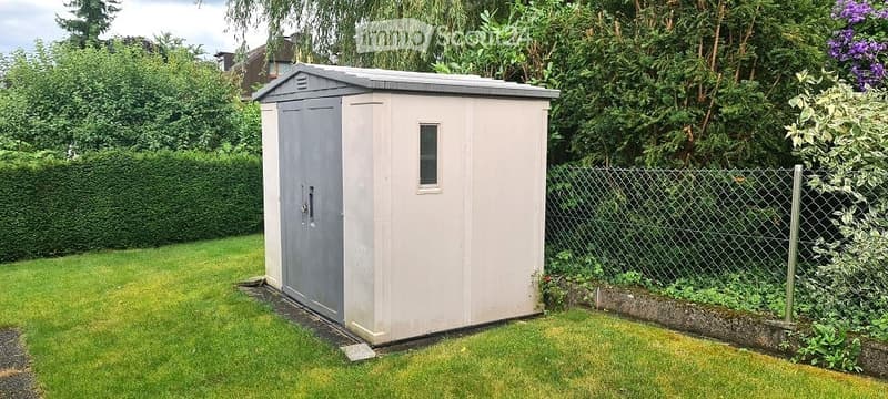 A small, single-story shed with a grey door and a small window, situated on a grassy lawn surrounded by a chain-link fence and lush greenery.