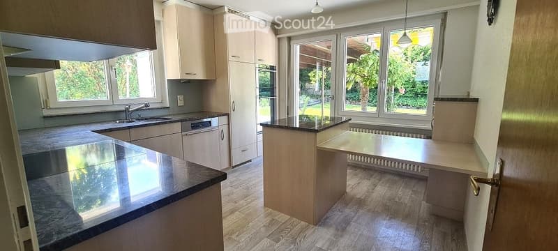 Modern kitchen with light wooden cabinets, granite countertops, island, and large windows overlooking a garden.