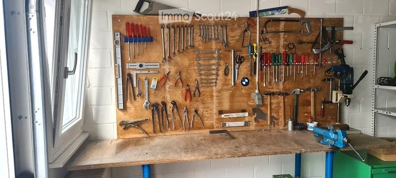 A well-organized workshop with a large wooden workbench, a pegboard wall displaying an array of tools including hammers, wrenches, screwdrivers, pliers, and a drill. The space is clean and functional, with a window on the left side providing natural light