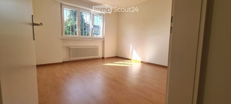 Empty room with wooden flooring, white walls, large window with radiator below, and a door visible on the left side.