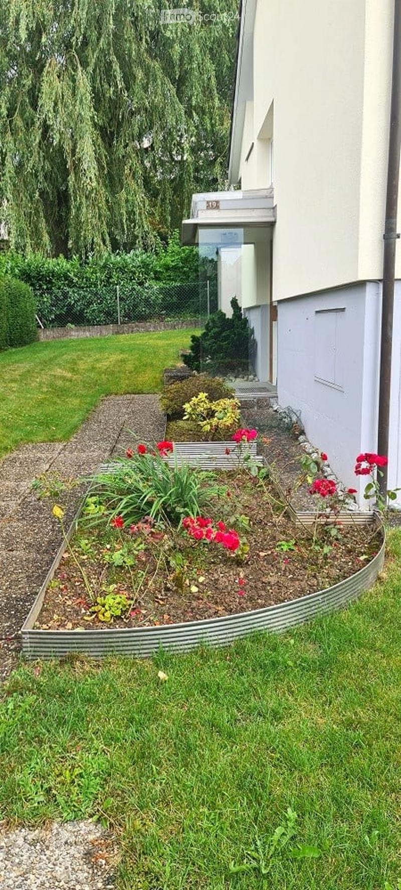 Well-maintained garden area with a variety of plants and flowers, including red and yellow blooms, neatly arranged in raised beds and containers. The garden features a gravel pathway and is bordered by a modern building with a white facade and a balcony.