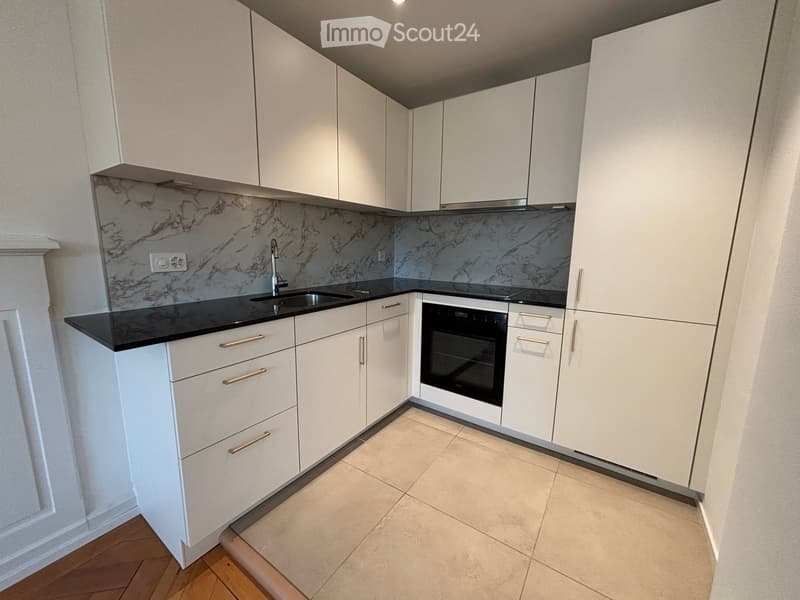 Modern kitchen with white cabinets, black countertops, and a marble backsplash. Features a sink, oven, and tiled flooring.