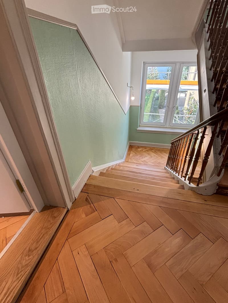 Spacious hallway with wooden herringbone flooring, light green walls, and a staircase with wooden balustrade leading upwards. A window on the right side allows natural light to enter.