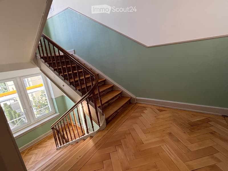 Spiral staircase with wooden steps and handrail, light green and white walls, herringbone wooden flooring, large window with view of greenery.
