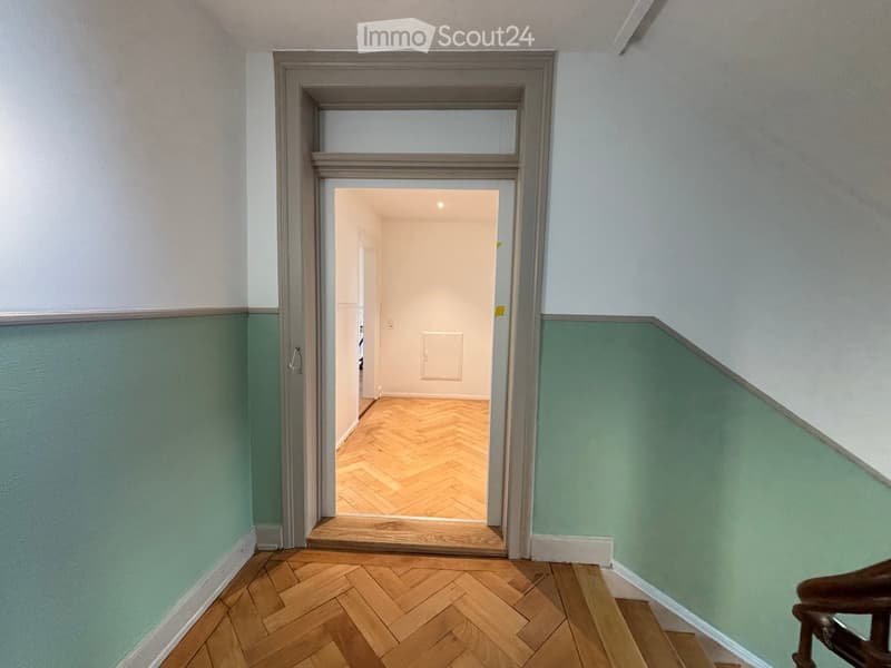 A narrow hallway with wooden herringbone flooring, light green walls, and a door leading to another room with white walls and a visible light fixture.