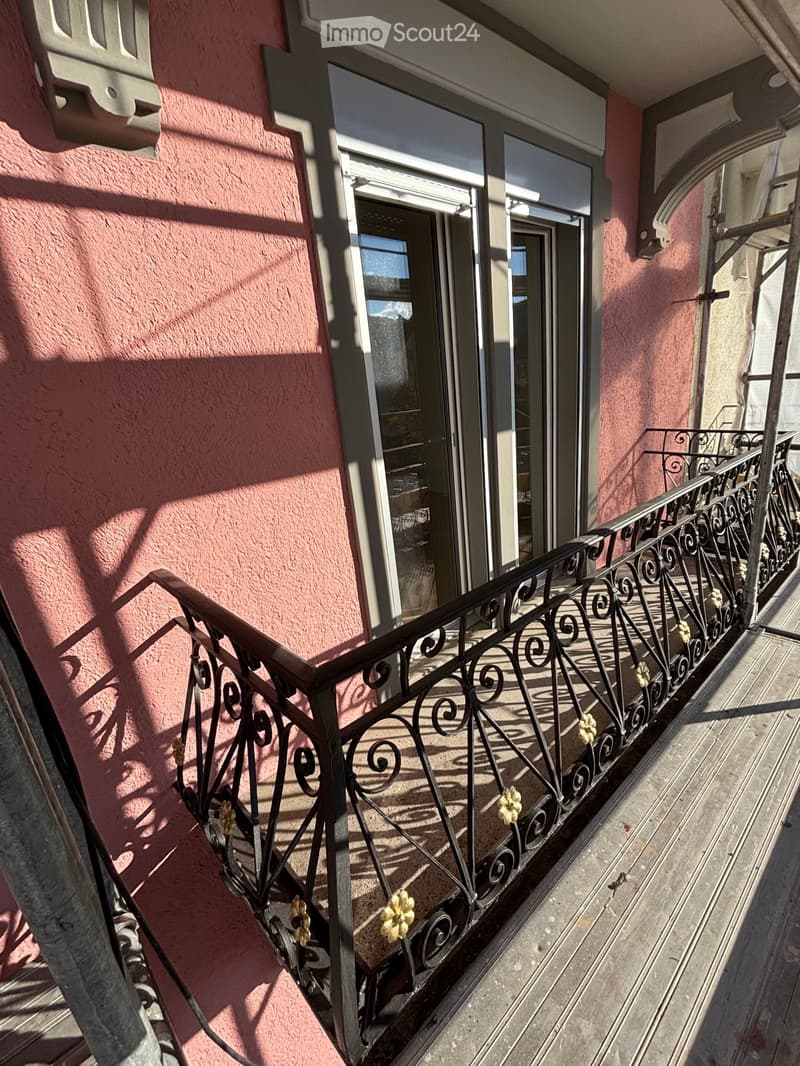 Balcony with ornate metal railing, pink building facade, and glass door leading to interior.