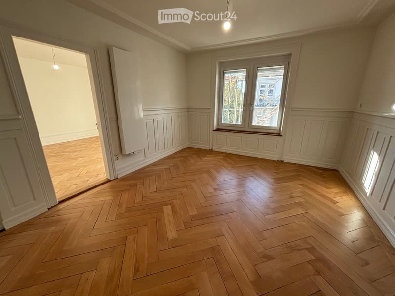 Empty room with herringbone wooden flooring, white walls, and large windows with a view of a building and greenery outside.