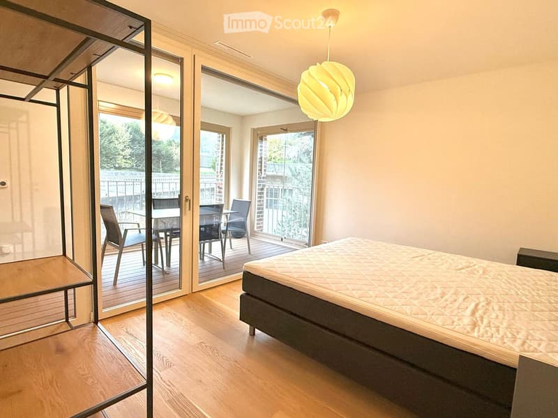 Modern bedroom with wooden flooring, large bed, pendant light, and sliding glass doors leading to a balcony with outdoor furniture.