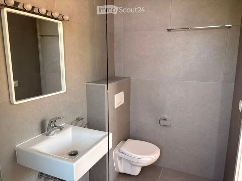 Modern bathroom with a white sink, chrome faucet, large mirror with built-in lighting, and a toilet. The walls are tiled in a neutral gray color.