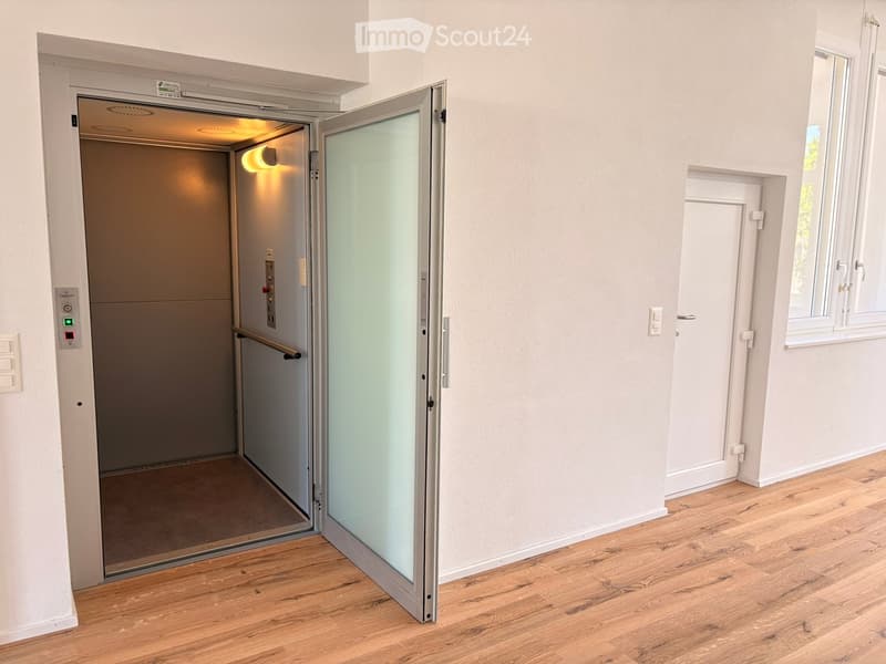 A modern hallway with wooden flooring, a glass-paneled elevator door, and a small window on the right side.