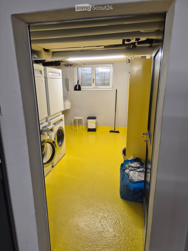 Laundry room with yellow flooring, washing machine, stool, trash bin, broom, and laundry basket.