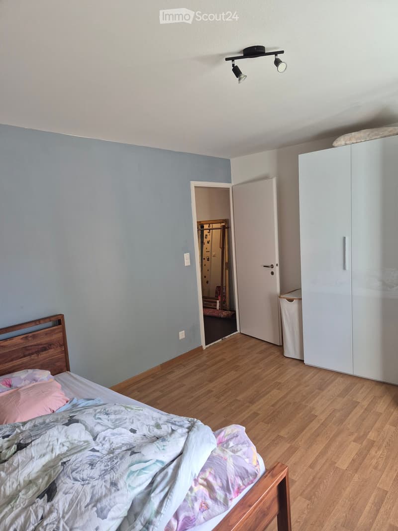 Bedroom with wooden floor, bed with colorful bedding, white wardrobe, and an open door leading to another room.
