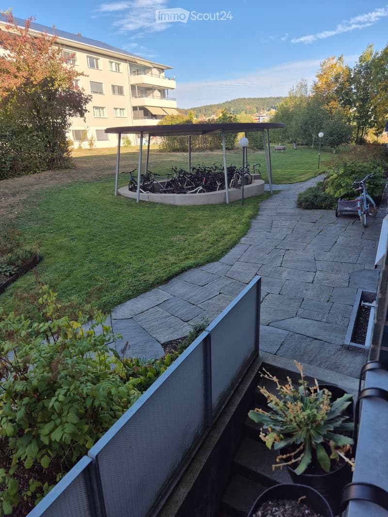 Balcony with potted plants, stone steps, metal railing, and a view of a grassy area with bicycles and a covered shelter.