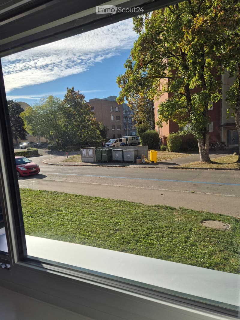 View from a balcony showing a street with parked cars, trash bins, and buildings in the background.