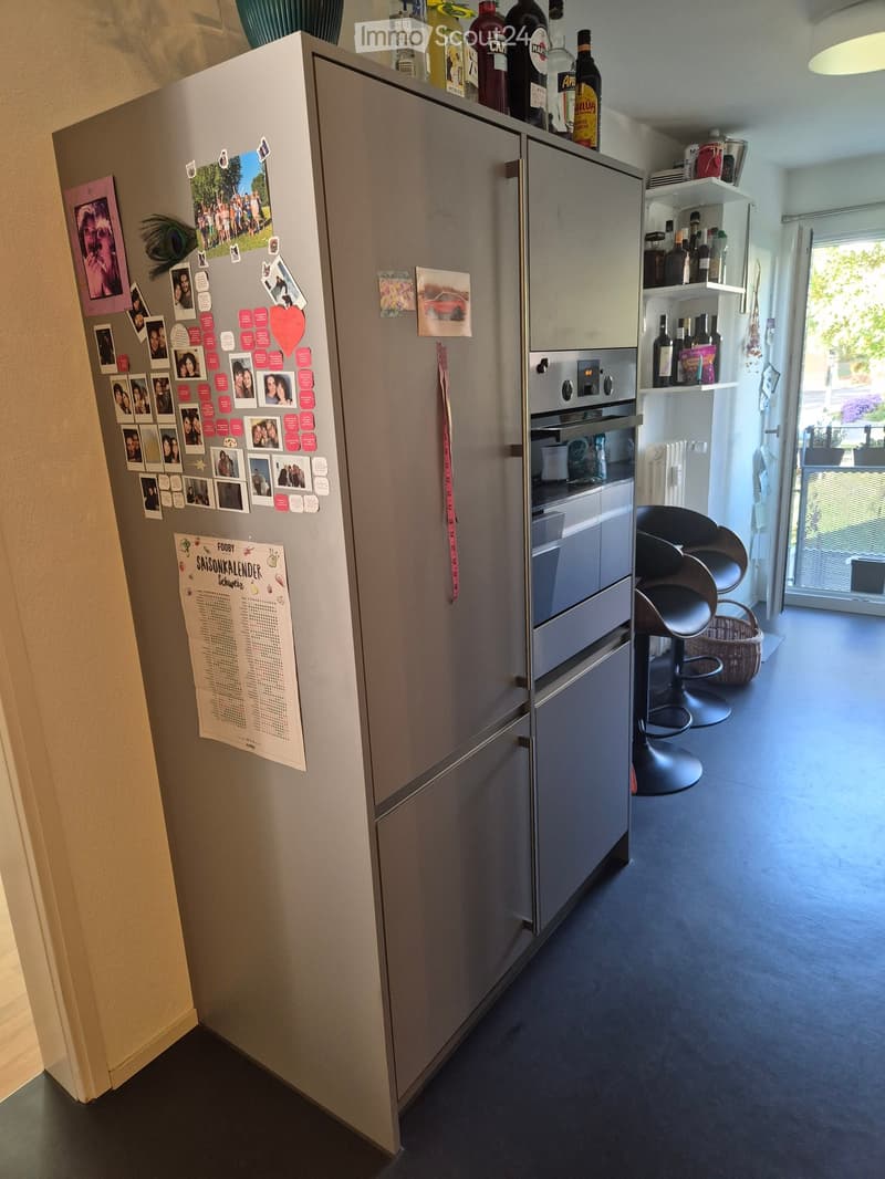 Modern kitchen with stainless steel refrigerator, oven, and bar stools. Shelves hold various bottles and items, with a calendar on the refrigerator.