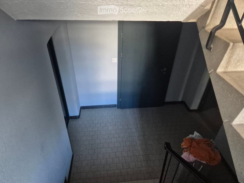 A narrow hallway with tiled floor, light blue walls, and a closed door at the end. A metal railing is visible on the right side, and some clothes are placed near the railing.