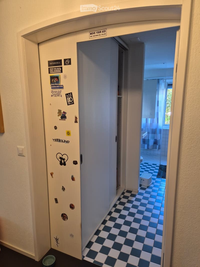 A narrow hallway with checkered floor tiles, leading to a bathroom with blue tiles and a small stool. The hallway walls are adorned with various stickers and signs, including one that reads "MOVE YOUR ASS AND YOUR MIND WILL FOLLOW".