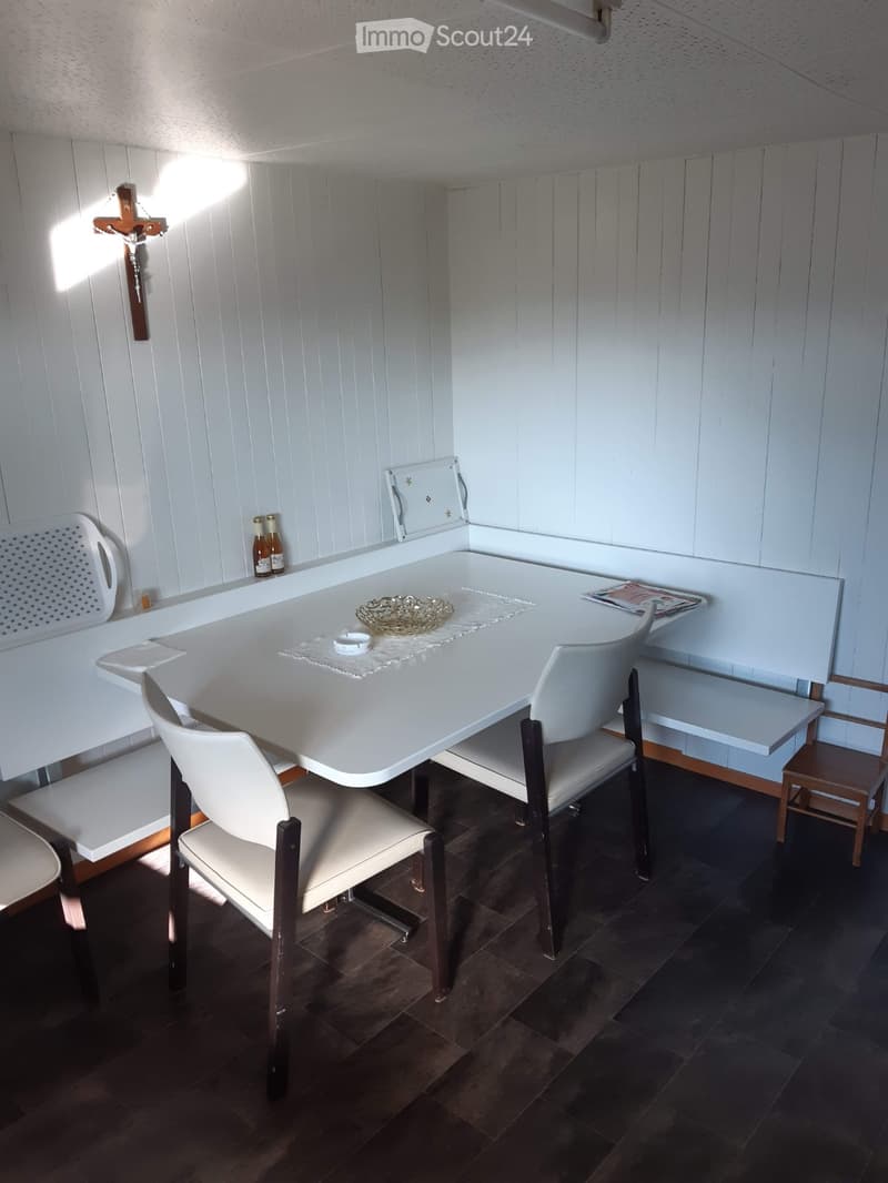 A small, furnished room with a white table, four chairs, a small stool, a cross on the wall, and two bottles on a shelf.
