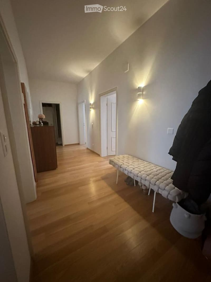 A narrow hallway with wooden flooring, white walls, and built-in lighting. A small bench sits against one wall, and a coat rack is visible on the right side.