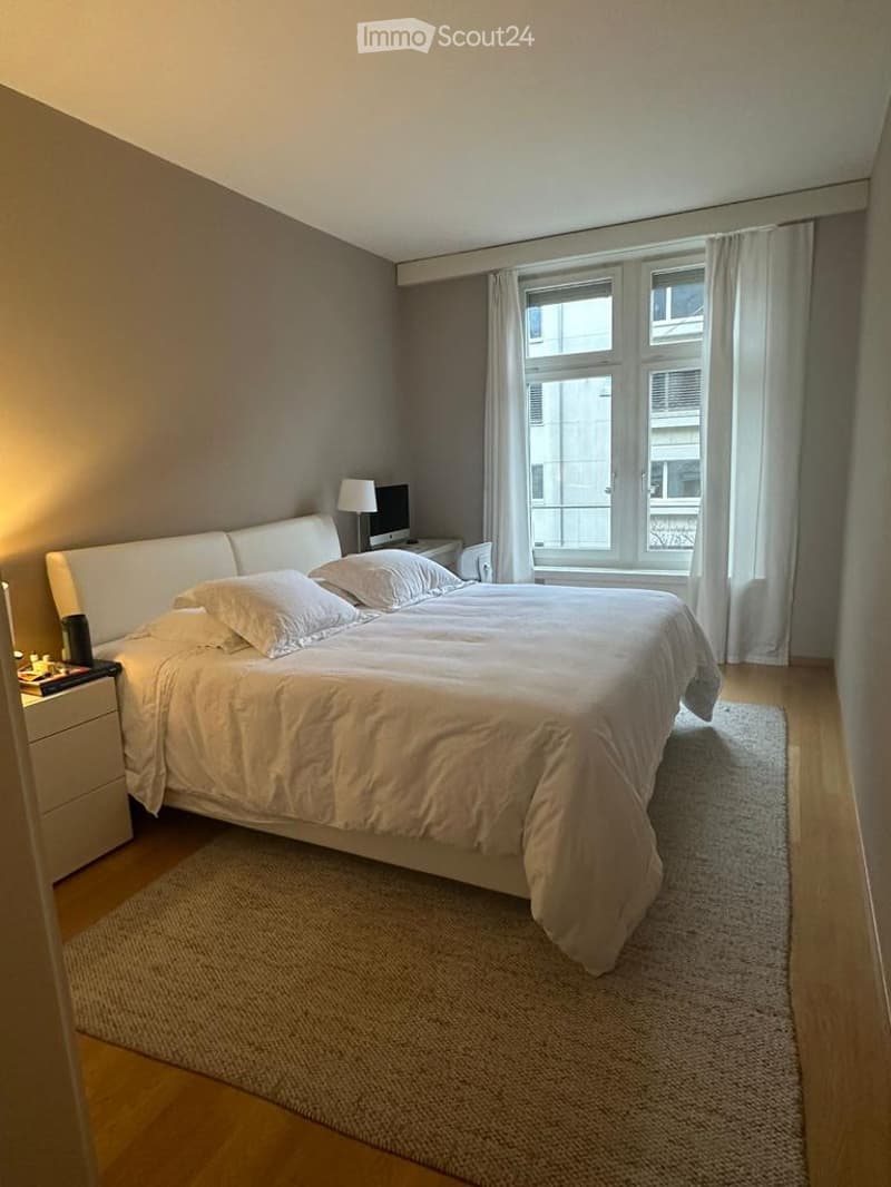 Modern bedroom with a large bed, nightstand, lamp, computer, and curtains on the window.