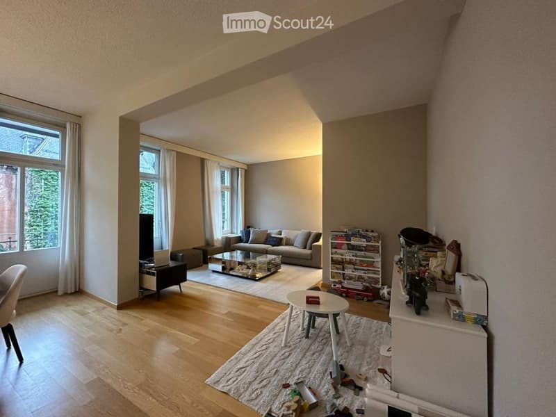 Bright living room with wooden flooring, large windows, a grey sofa, coffee table, small round table, bookshelf, and various decorative items.