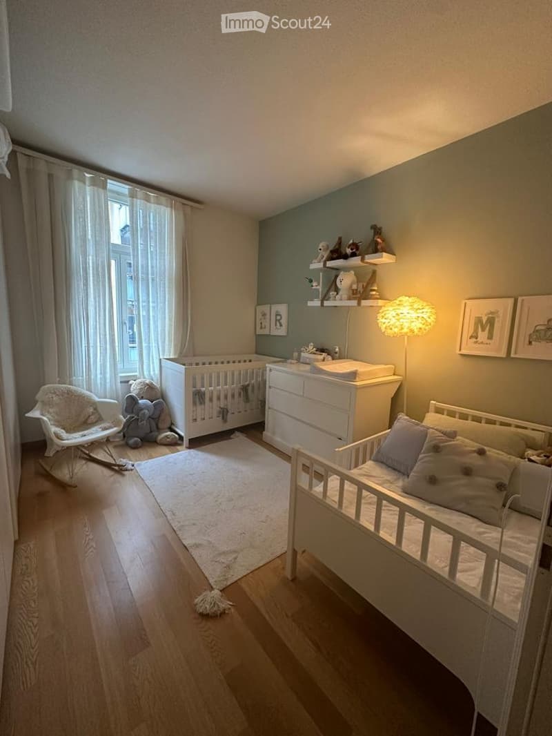 A well-lit, modern nursery with a crib, daybed, rocking chair, changing table, shelves with toys, and picture frames on the walls.