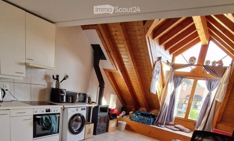 A cozy attic space with exposed wooden beams, a kitchen area with cabinets, stove, microwave, and washing machine, and a seating area with a stool and table.