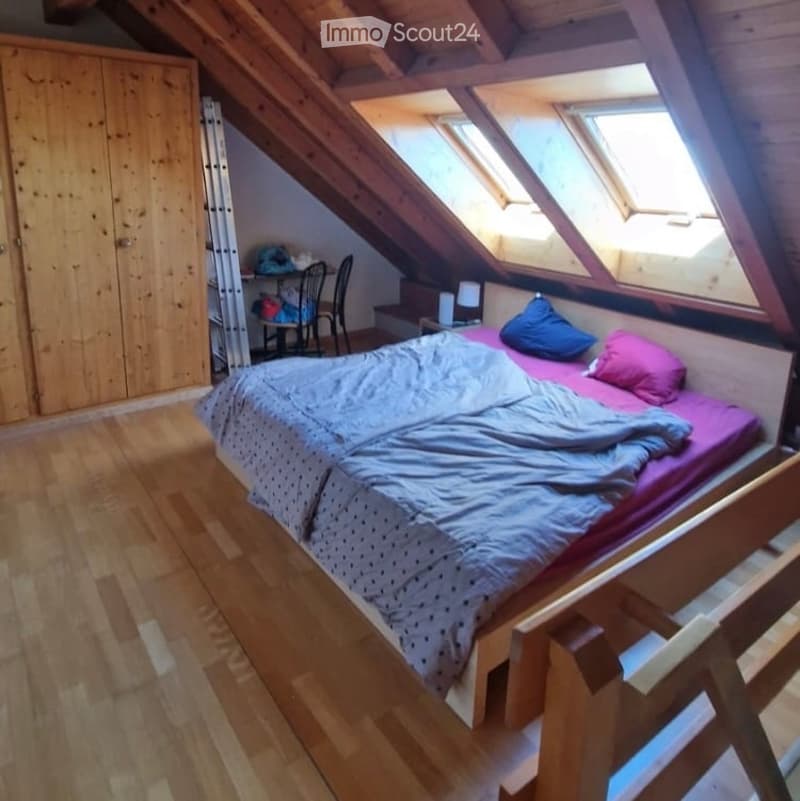 Cozy attic bedroom with wooden floors, slanted wooden ceiling, skylights, a bed with pillows and blankets, a small table with a lamp, a chair, and a wooden cabinet.