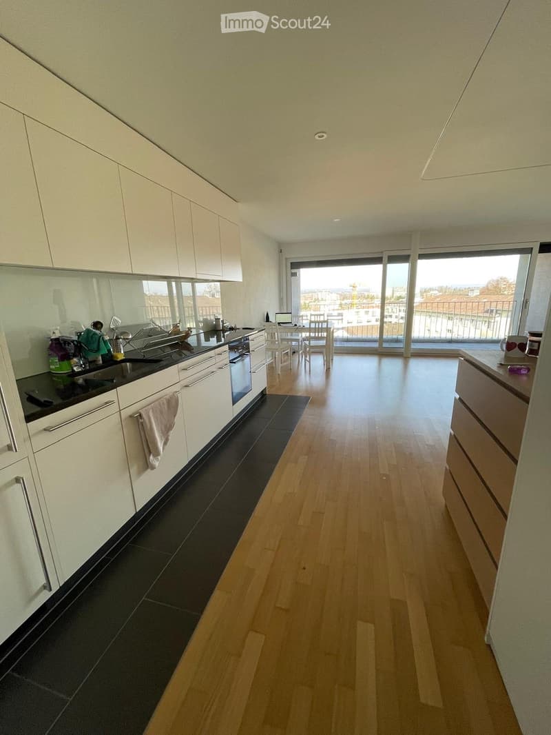 Modern kitchen with white cabinets, black countertop, and wooden flooring. Features an oven, sink, dish rack, and a small dining area with a table and chairs.