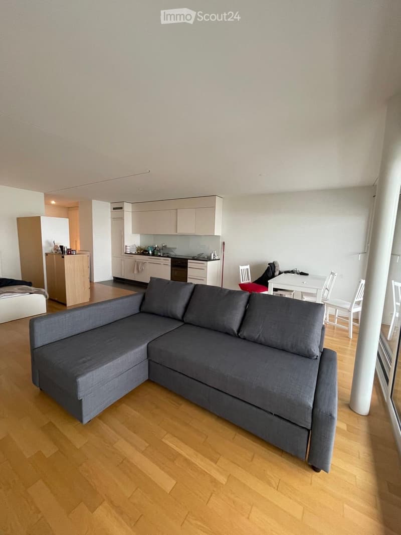A modern, open-concept living space with a grey sectional sofa, a small dining table with chairs, and a compact kitchen area featuring light-colored cabinets, an oven, and a refrigerator.