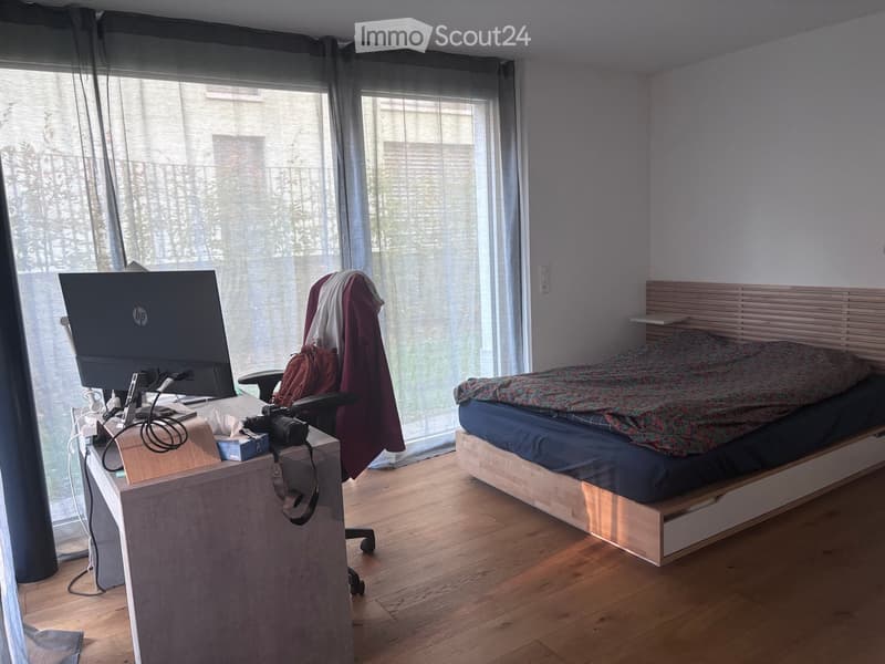A modern bedroom with a wooden bed, a laptop on a desk, a chair, and a camera on the desk. The room has large windows with sheer curtains and wooden flooring.