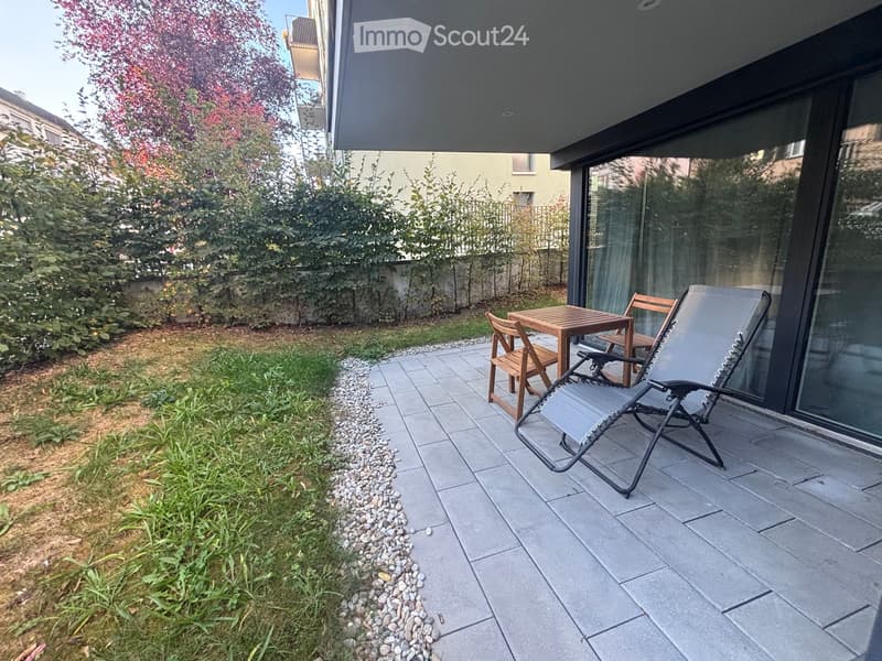 Outdoor patio with tiled floor, small wooden table, two chairs, and a lounge chair, surrounded by greenery and a stone border.