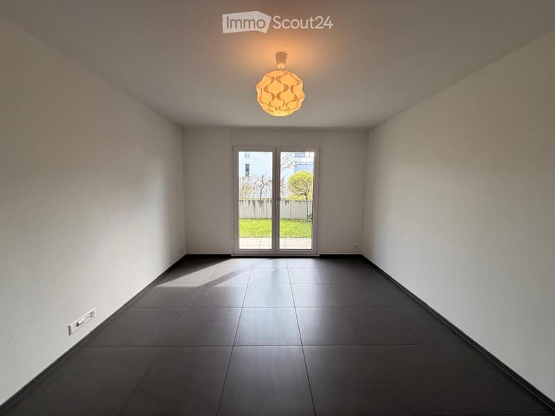 Empty room with dark tiled floor, white walls, and a large sliding glass door leading to a garden area.