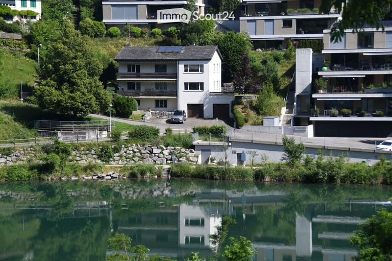 A modern two-story house with a garage, situated on a hillside overlooking a calm river. The house features large windows, a flat roof, and is surrounded by lush greenery. A car is parked in the driveway, and the house has a contemporary design with clean