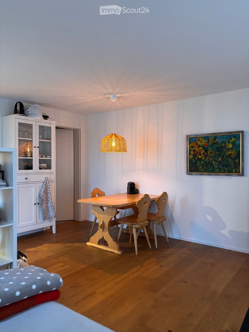Bright kitchen with wooden floor, white walls, a wooden dining table with four chairs, a white cabinet with glass doors, a hanging lamp, and a painting on the wall.