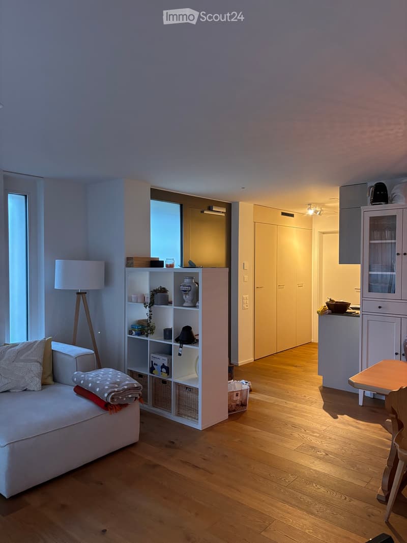 A modern, well-lit living room with a white sofa, a wooden table, a shelf with decorative items, and a lamp. The room features light wooden flooring and large windows allowing natural light to fill the space.