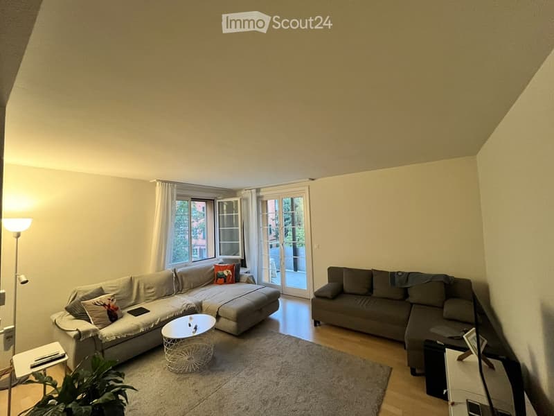 Modern living room with two sectional sofas, a white coffee table, a small side table, a floor lamp, and a plant near the window. The room features large windows and sliding glass doors leading to a balcony.